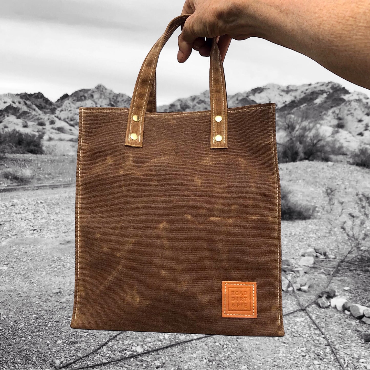 A brown waxed canvas tote bag held by the handles to show its small size. The bag has light brown, short straps. There is white stitching along the edges and brass rivets.