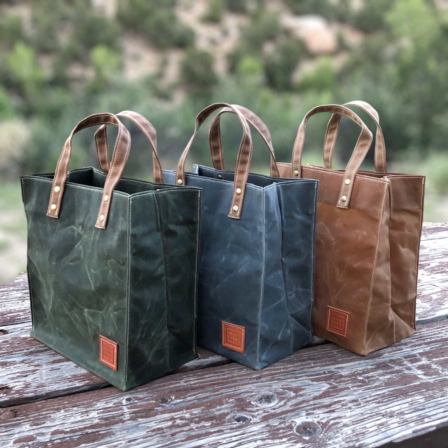 Three waxed canvas tote bags in green, gray and brown. Each bag has short brown handles with brass rivets, and a small square leather patch.