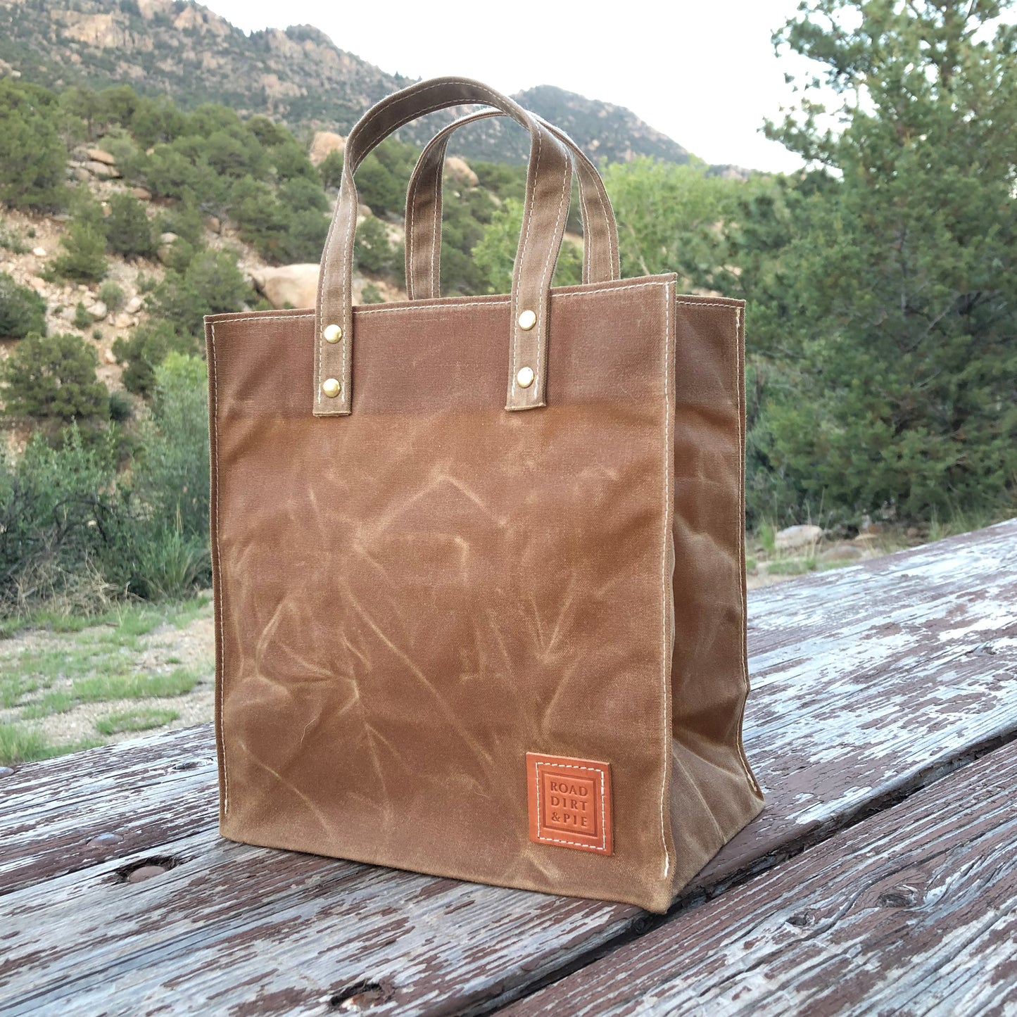 A brown canvas tote bag with light brown short handles held secure with brass rivets. The bag is standing upright and has white stitches on the edges and a small leather patch.